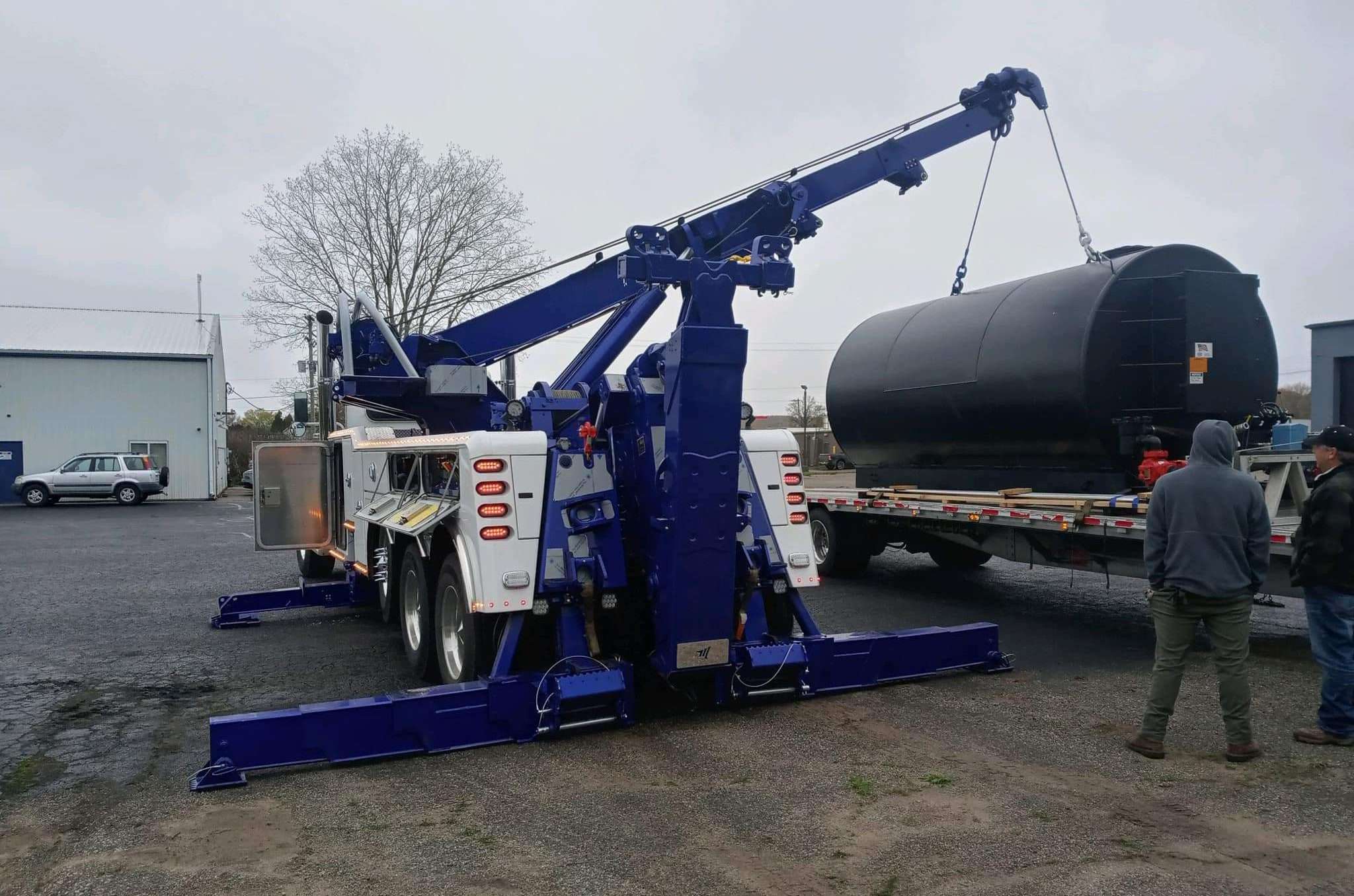 A heavy-duty tow truck using its rotating boom to move a large oil tank.